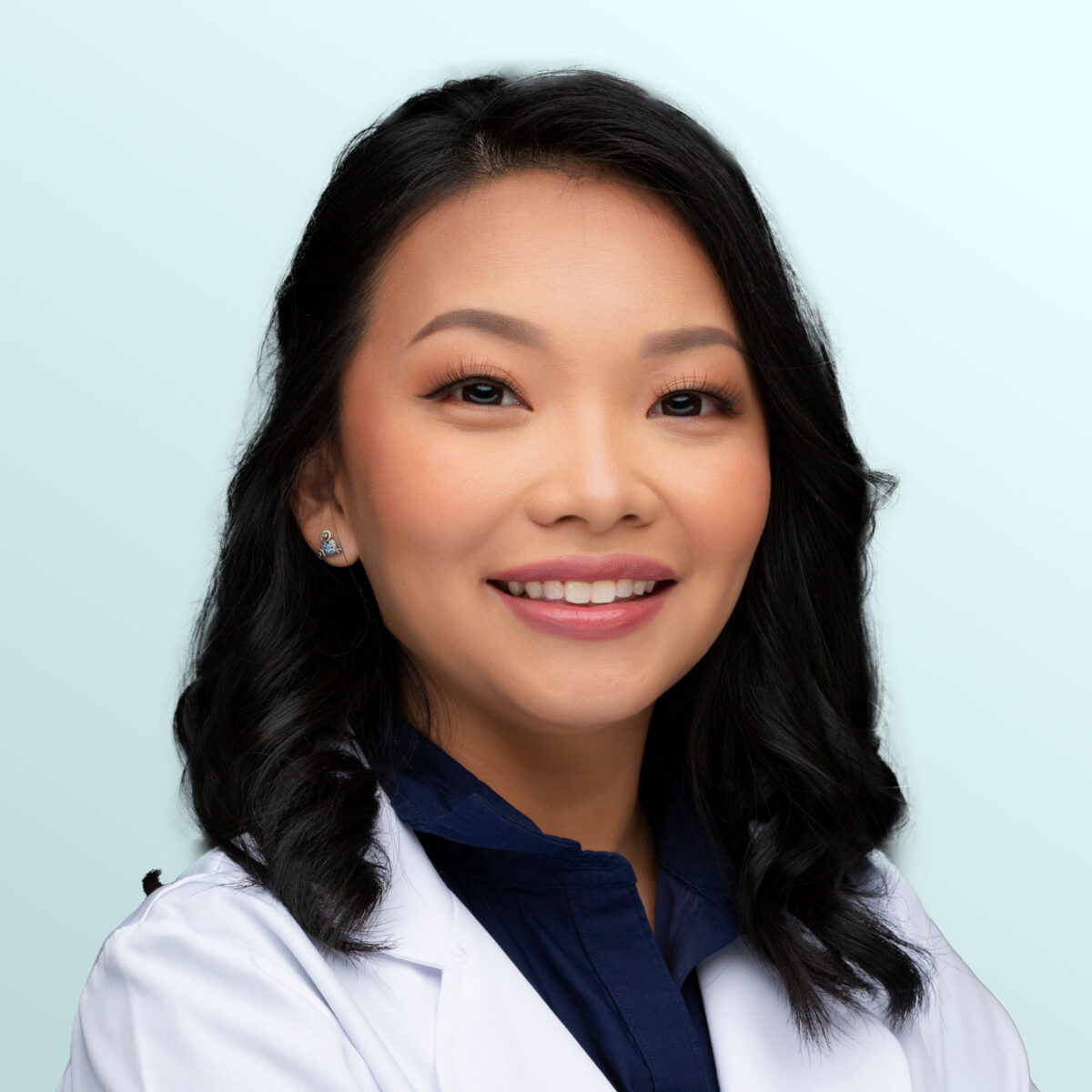 A woman with wavy black hair wearing a white lab coat and navy blouse smiles at the camera against a light blue background.