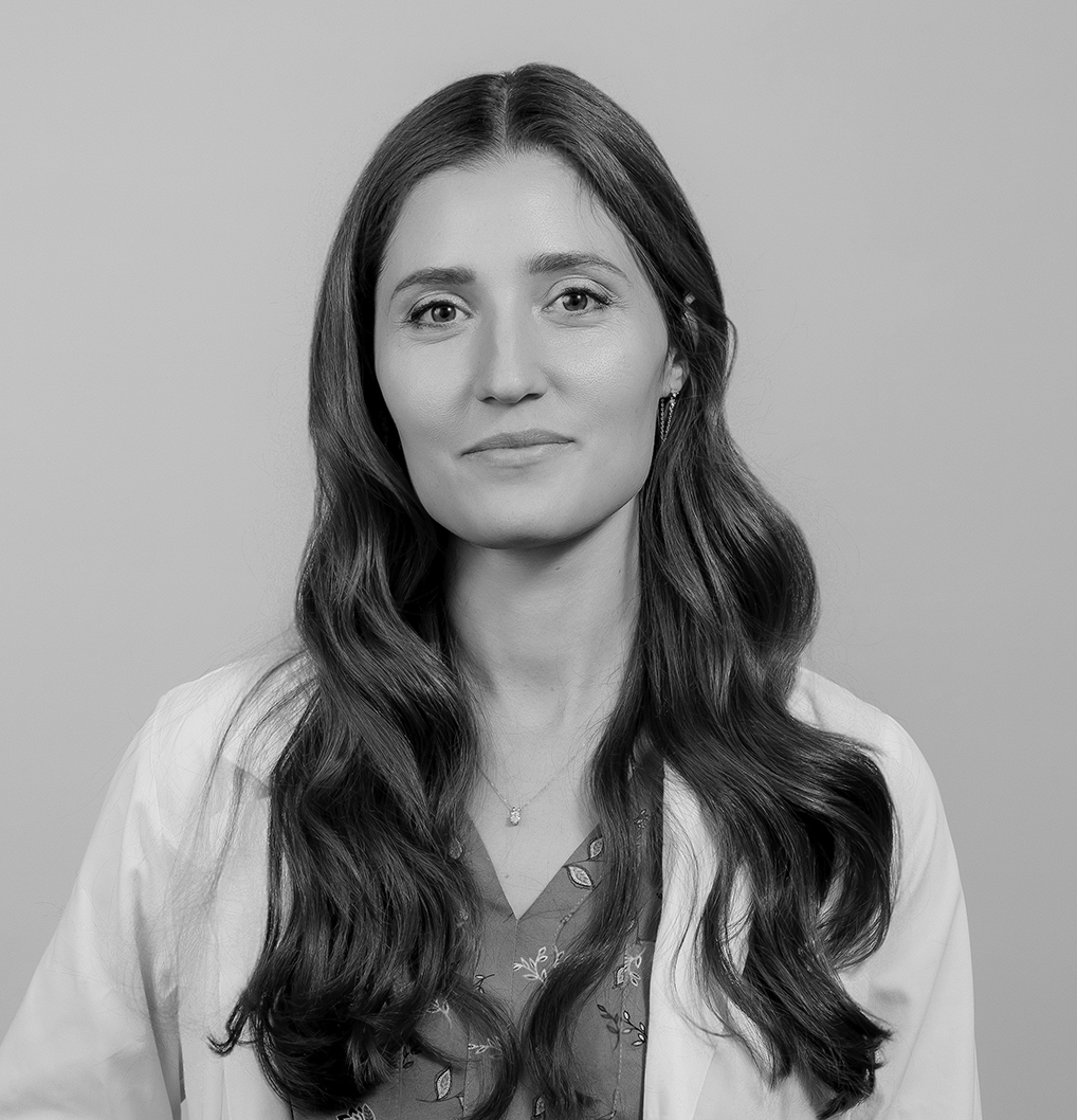 Black and white portrait of a woman with long wavy hair, wearing a light-colored lab coat over a patterned blouse, looking at the camera with a neutral expression against a plain background.
