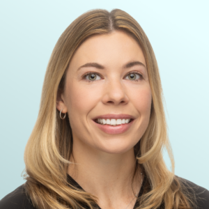 A woman with long, light brown hair and hazel eyes smiles at the camera. She is wearing a grey top and small earrings, with a light blue background behind her.