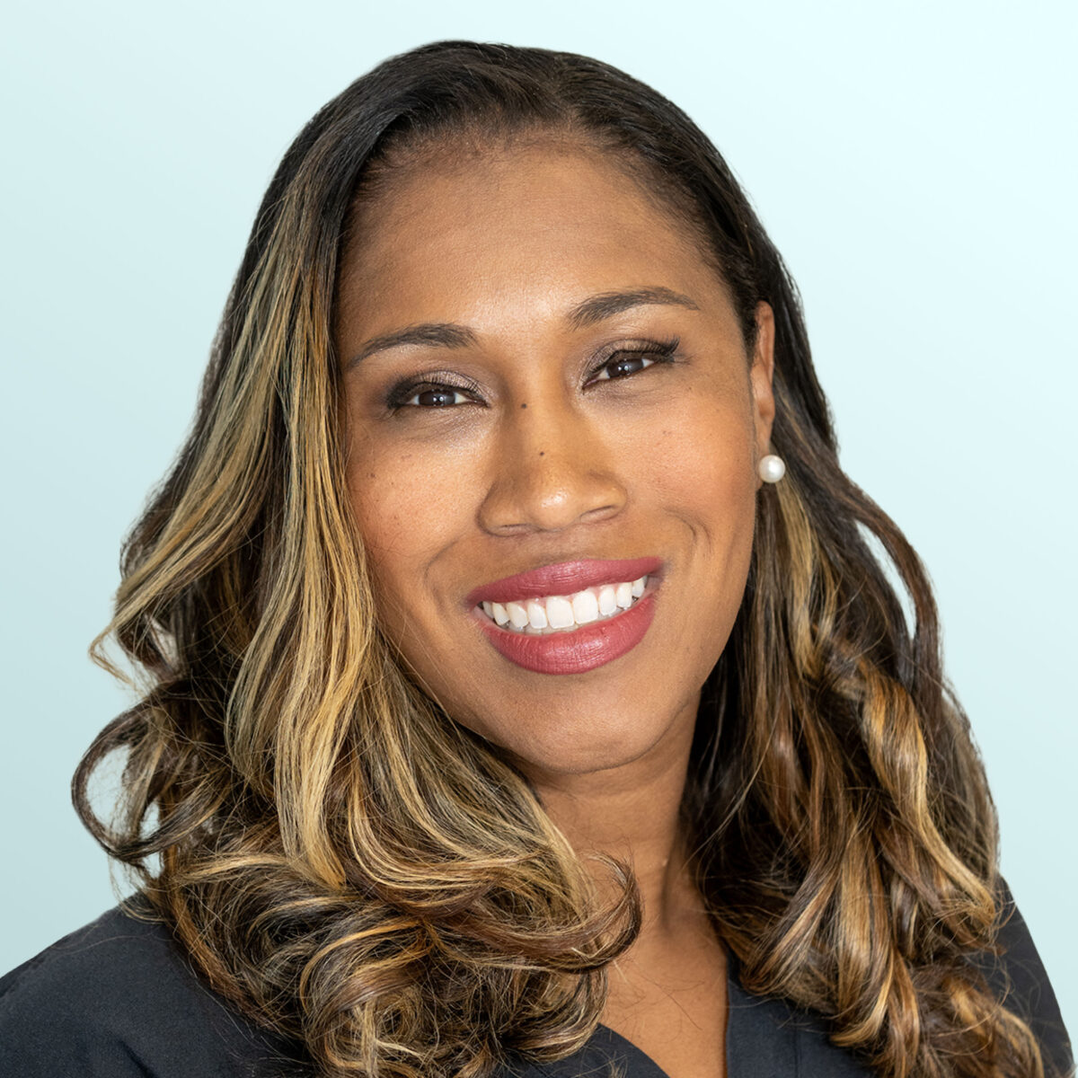 A woman with long, wavy, highlighted hair smiles at the camera. She is wearing pearl earrings and a dark scrub top with embroidered text, standing against a light blue background.