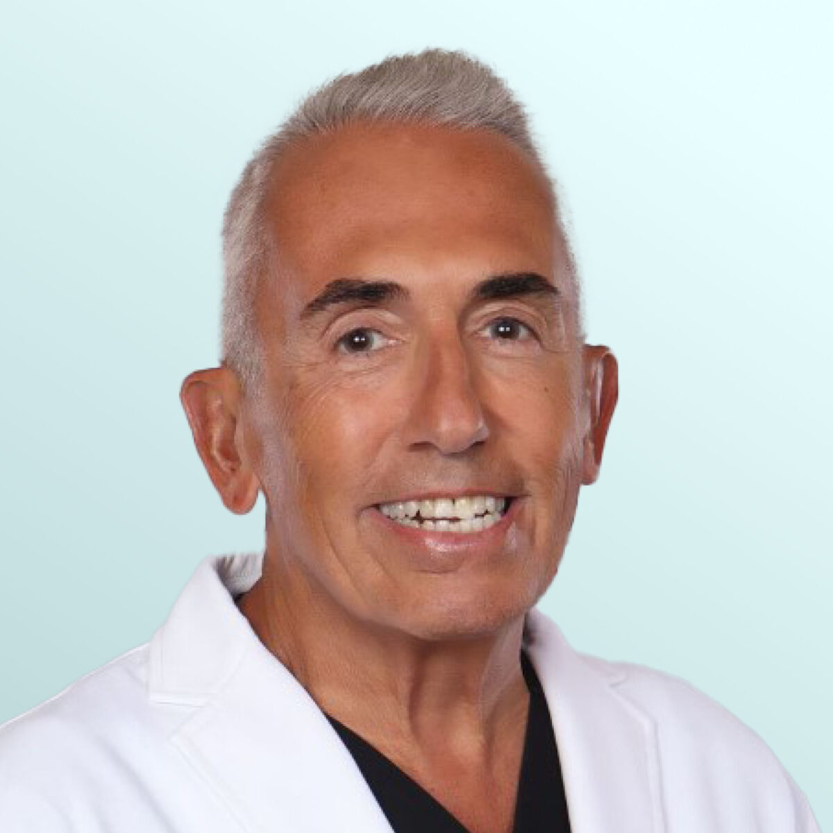 Smiling older man with short gray hair wearing a white lab coat and black shirt, posed against a light blue background.