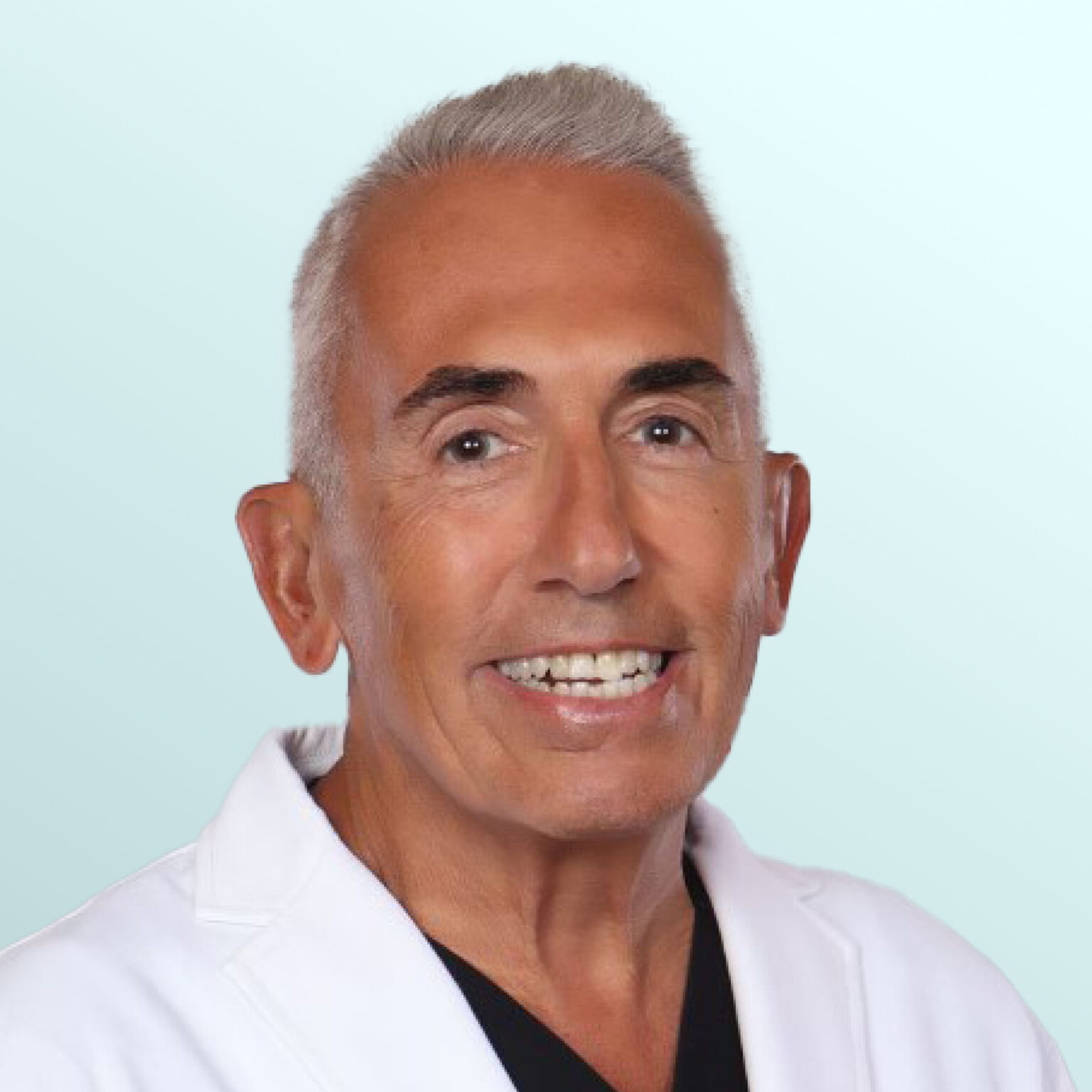 Smiling older man with short gray hair wearing a white lab coat and black shirt, posed against a light blue background.