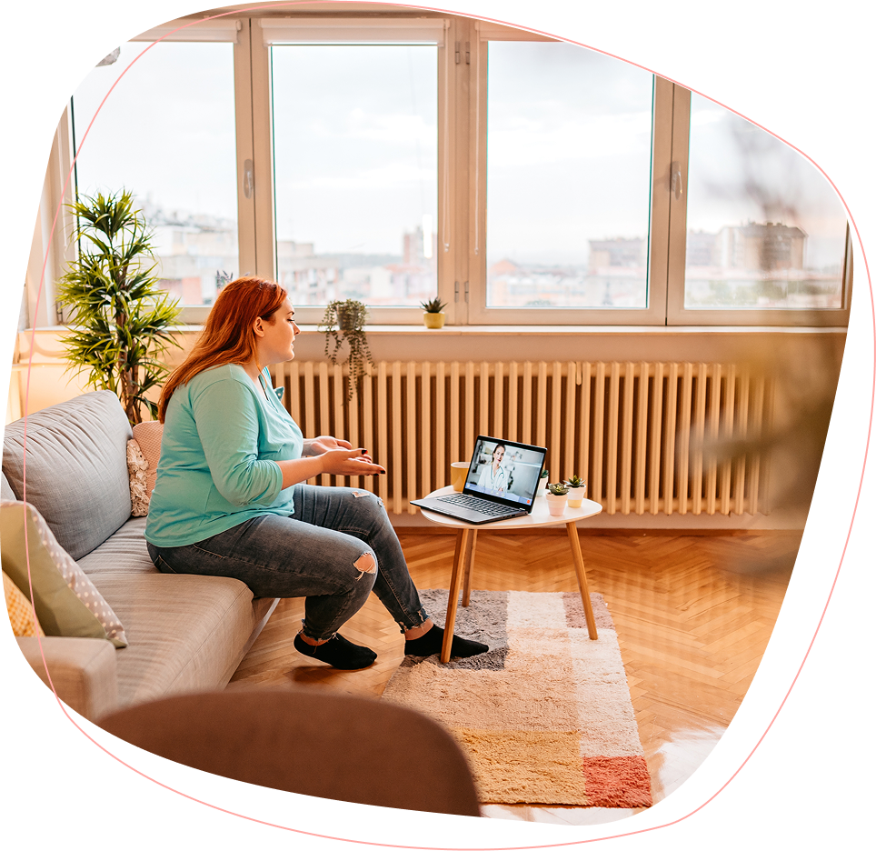 A woman sits on a sofa in a cozy living room, having a video call on her laptop placed on a small table. She gestures with her hands while talking, with large windows and city buildings visible in the background.