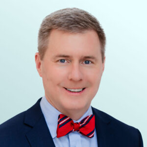 A man in a navy suit with a light blue shirt and a red striped bow tie smiles at the camera against a light blue background.