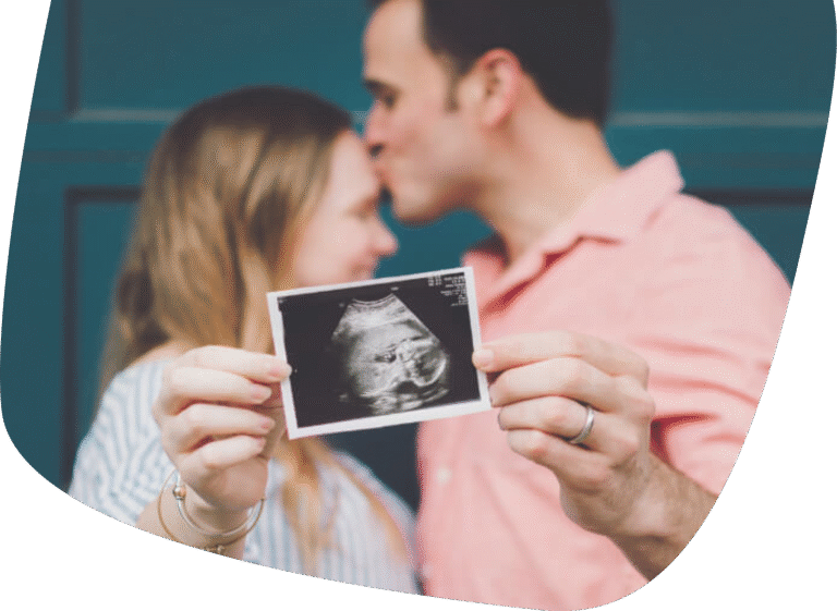 A couple holds up an ultrasound photo, with the man kissing the womans forehead. The focus is on the ultrasound image in the foreground, while the couple is blurred in the background.