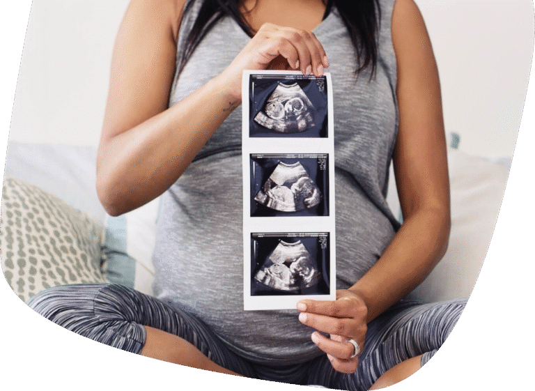 A pregnant person in grey athletic wear sits cross-legged on a bed, holding up three ultrasound images showing their developing baby.