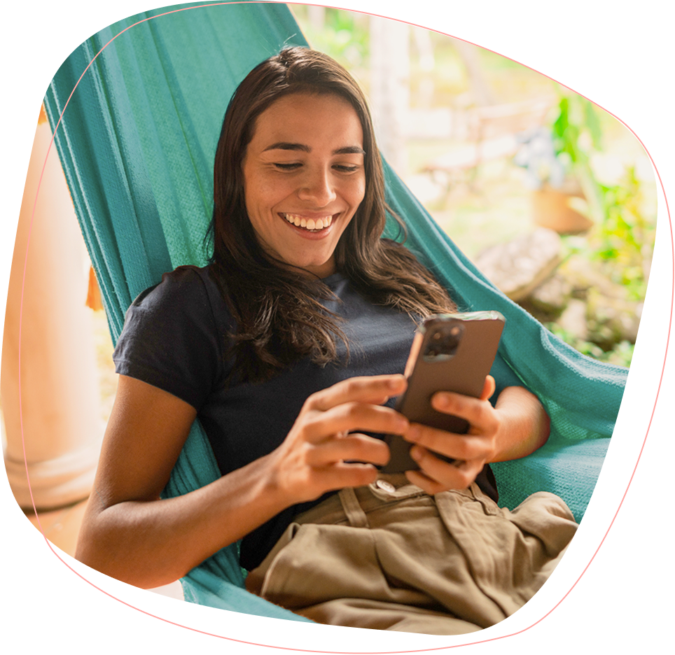 A smiling person with long dark hair relaxes in a turquoise hammock while looking at and using a smartphone. They are wearing a dark t-shirt and beige pants, and appear to be outdoors.