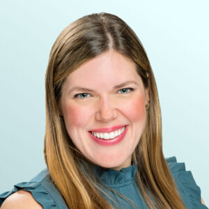 A woman with long, straight light brown hair smiles at the camera. She is wearing a ruffled, sleeveless teal blouse and is posed against a light blue background.