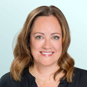 Smiling woman with shoulder-length light brown hair, wearing a black blouse and gold necklaces, posing in front of a light blue background.