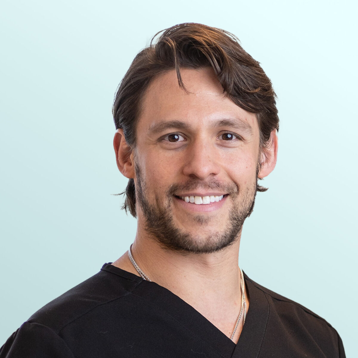 A smiling man with medium-length brown hair and a short beard, wearing a black V-neck shirt and a silver necklace, stands in front of a light blue background.