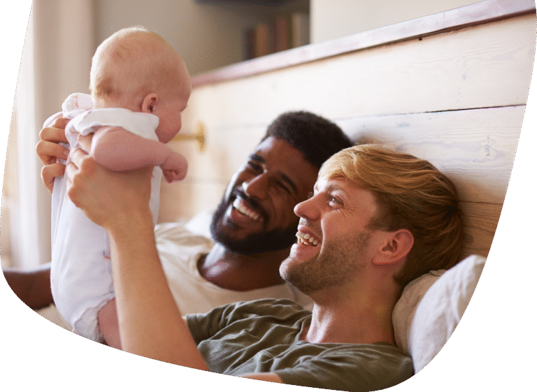 Two men lying on a bed smile and laugh while one holds up a baby, who looks back at them. The setting is warm and relaxed, suggesting a happy family moment.