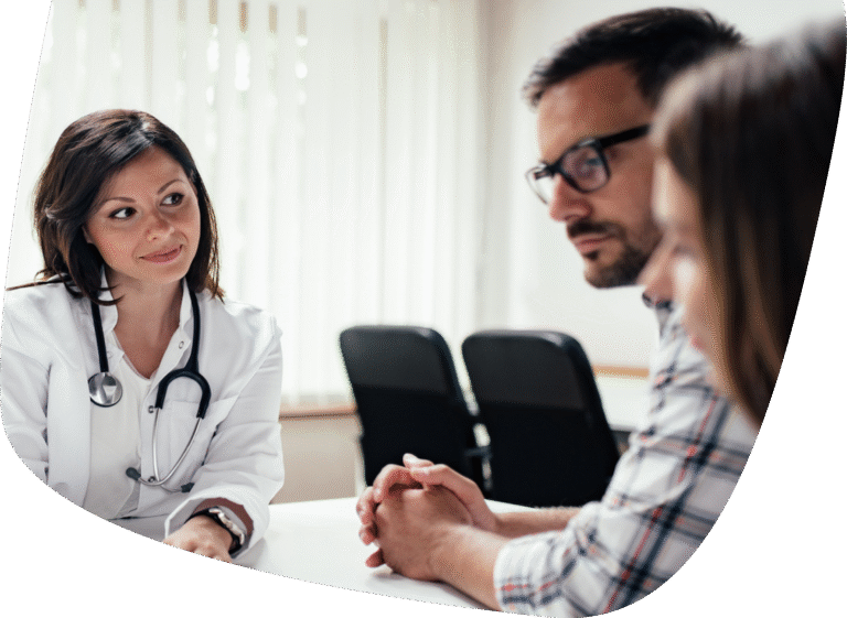 A female doctor wearing a white coat and stethoscope sits at a desk, attentively listening to a man and woman seated across from her, having a serious conversation in a medical office.
