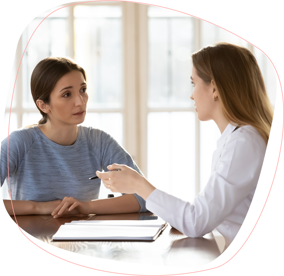 Two women sit across from each other at a table, engaged in a serious conversation. One woman listens attentively while the other, holding a pen, appears to be explaining something. A clipboard is on the table.