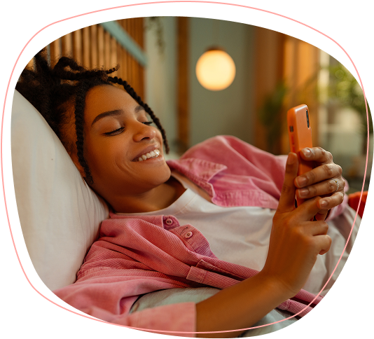 A young woman with braided hair smiles while lying on a couch, wearing a pink shirt and holding a smartphone in both hands. The background shows a warm, softly lit room.
