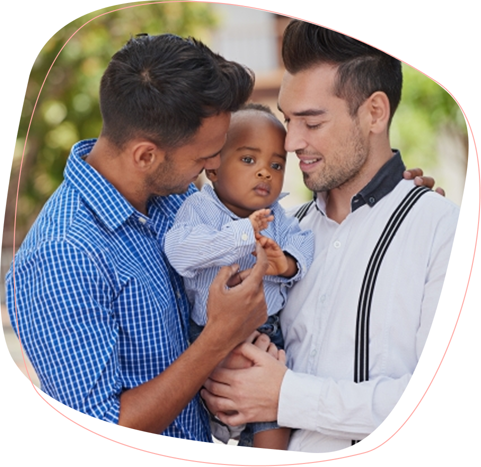 Two men lovingly hold a young child between them. All three are smiling and dressed in coordinated blue and white shirts. The background is softly blurred, suggesting an outdoor setting.