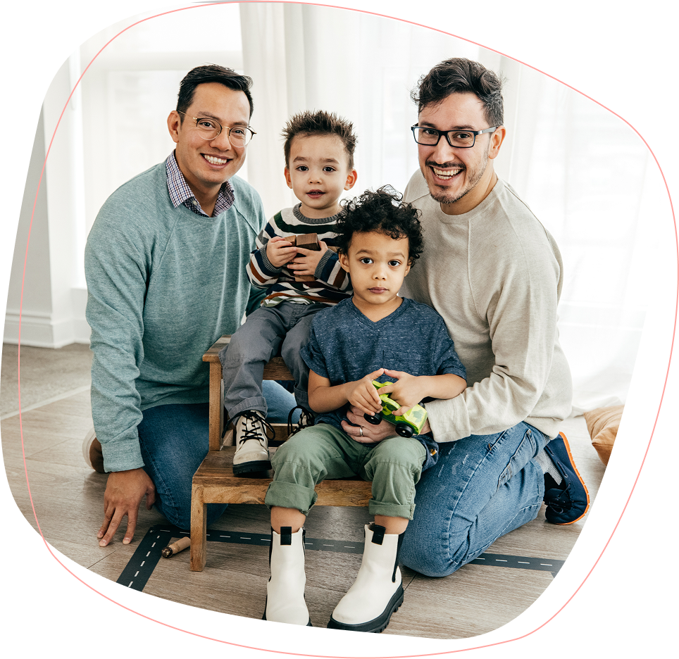 Two adults and two children pose together indoors. The adults are kneeling and smiling, while the children sit on a small chair holding toys. Natural light comes through windows behind them.