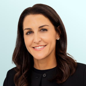 A woman with long brown hair and a black sleeveless top smiles at the camera against a light blue background. She is wearing gold jewelry, including a necklace and earrings.