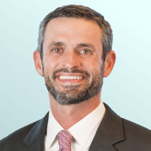 A man with short dark hair and a beard is smiling, wearing a dark suit jacket, white shirt, and a colorful patterned tie. The background is a light blue gradient.