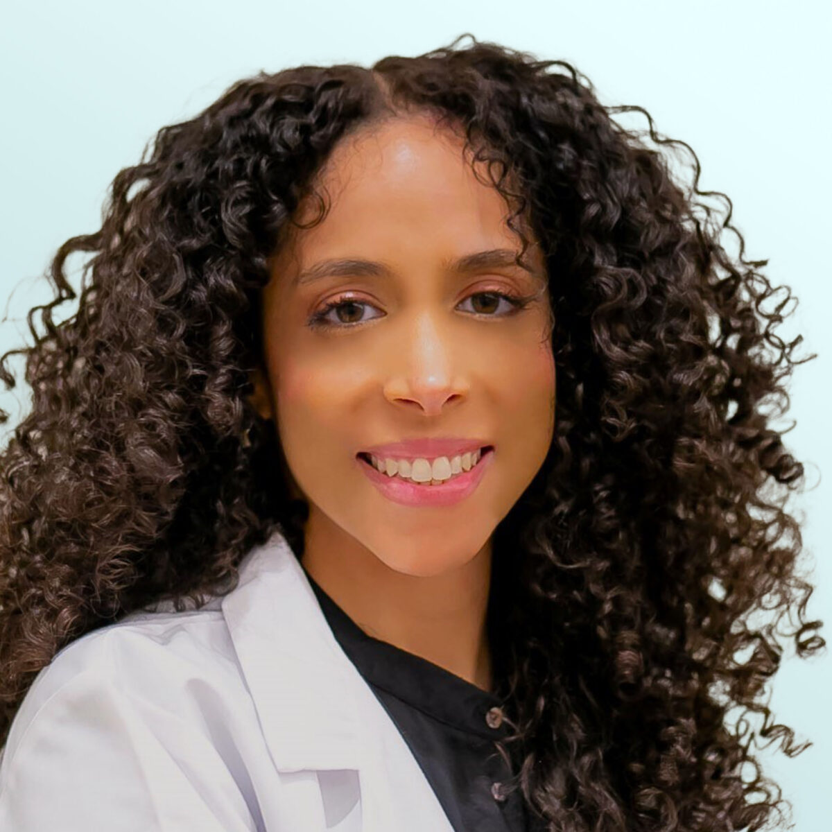 A woman with long, curly hair wears a white lab coat and a black shirt, smiling at the camera against a light blue background.