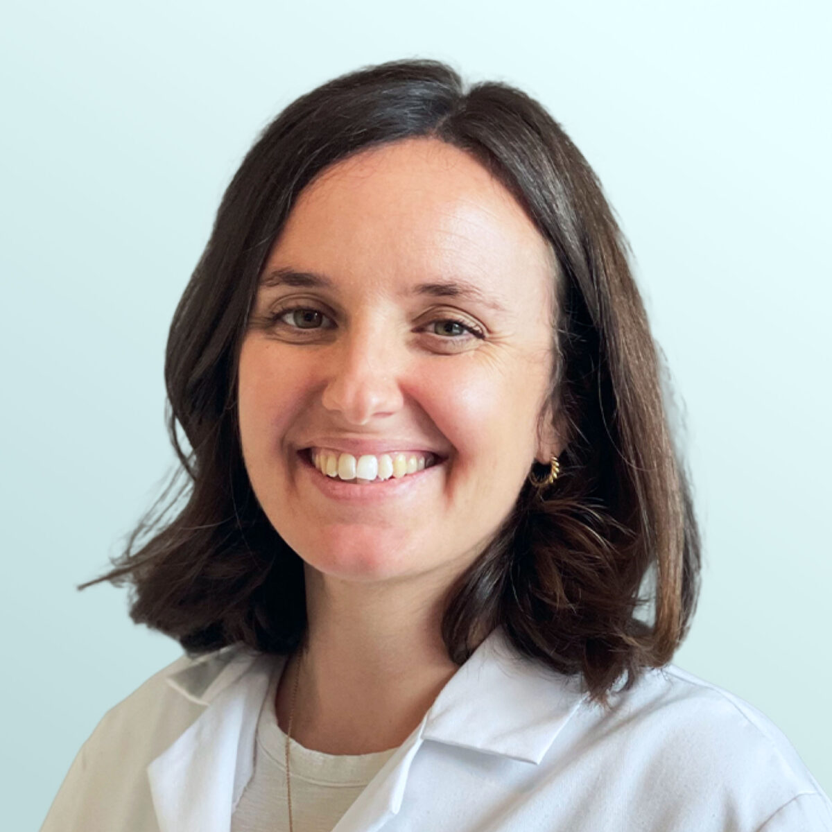 A woman with shoulder-length brown hair smiles at the camera, wearing a white lab coat and a light-colored top, standing against a plain light blue background.