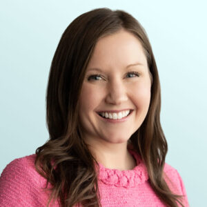 A woman with long brown hair, wearing a bright pink sweater, smiles at the camera against a light blue background.