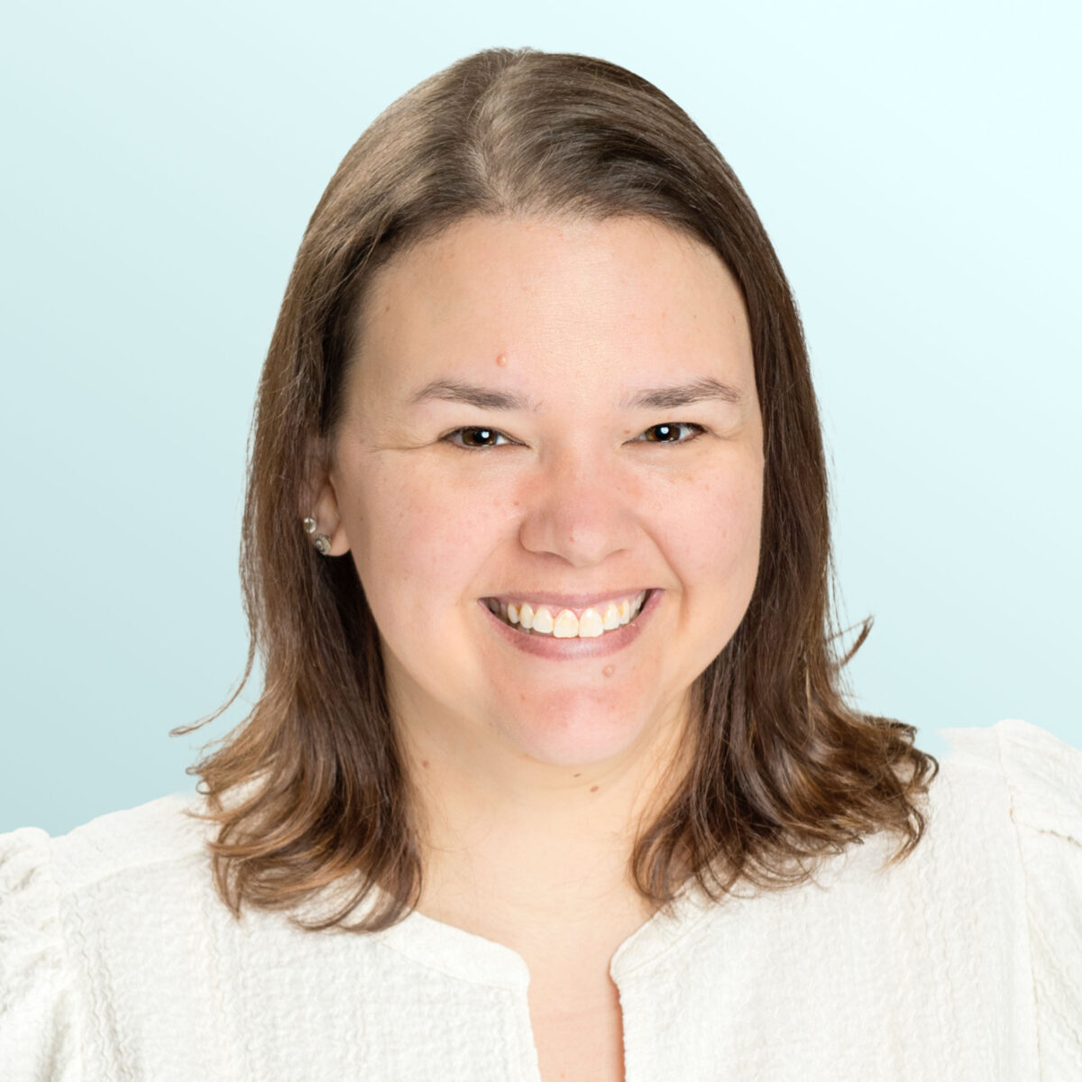 A woman with shoulder-length brown hair smiles at the camera. She is wearing a textured white top and is posed in front of a light blue background.
