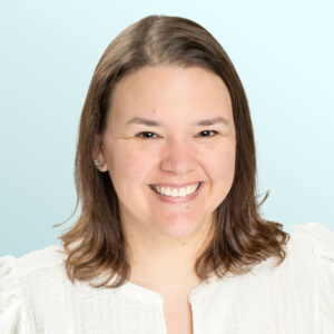 A woman with shoulder-length brown hair smiles at the camera. She is wearing a textured white top and is posed in front of a light blue background.