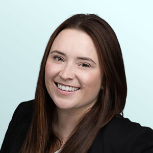 A woman with long straight brown hair, wearing a black blazer and white top, smiles at the camera against a light blue background.