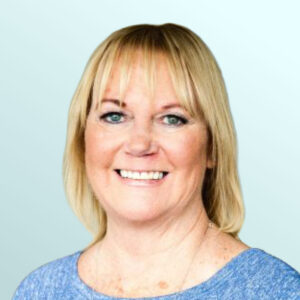 Smiling woman with blonde hair and bangs, wearing a blue top, standing in front of a light blue background.
