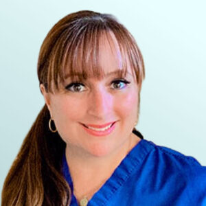 A woman with long brown hair in a ponytail, wearing blue medical scrubs, smiles at the camera against a light blue background.