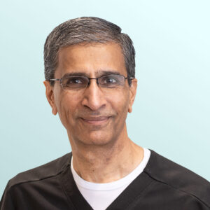 A middle-aged man with short gray hair and glasses, wearing a black shirt over a white undershirt, smiles slightly at the camera against a light blue background.