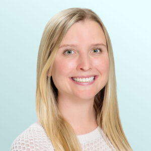 A smiling young woman with long straight blonde hair, wearing a white textured sweater, poses in front of a light blue background.