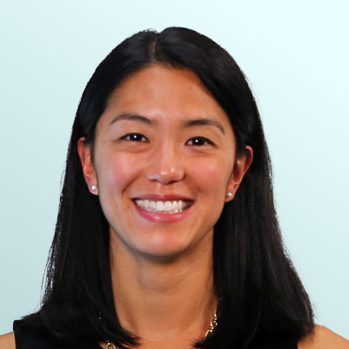 A woman with straight black hair and a floral necklace, wearing a sleeveless black top, smiles in front of a light blue background.
