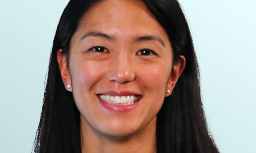 A woman with straight black hair and a floral necklace, wearing a sleeveless black top, smiles in front of a light blue background.