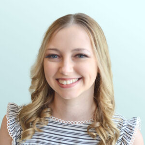 A young woman with long blonde hair and blue eyes smiles at the camera. She is wearing a black and white striped top with ruffled sleeves, and the background is a solid light blue color.