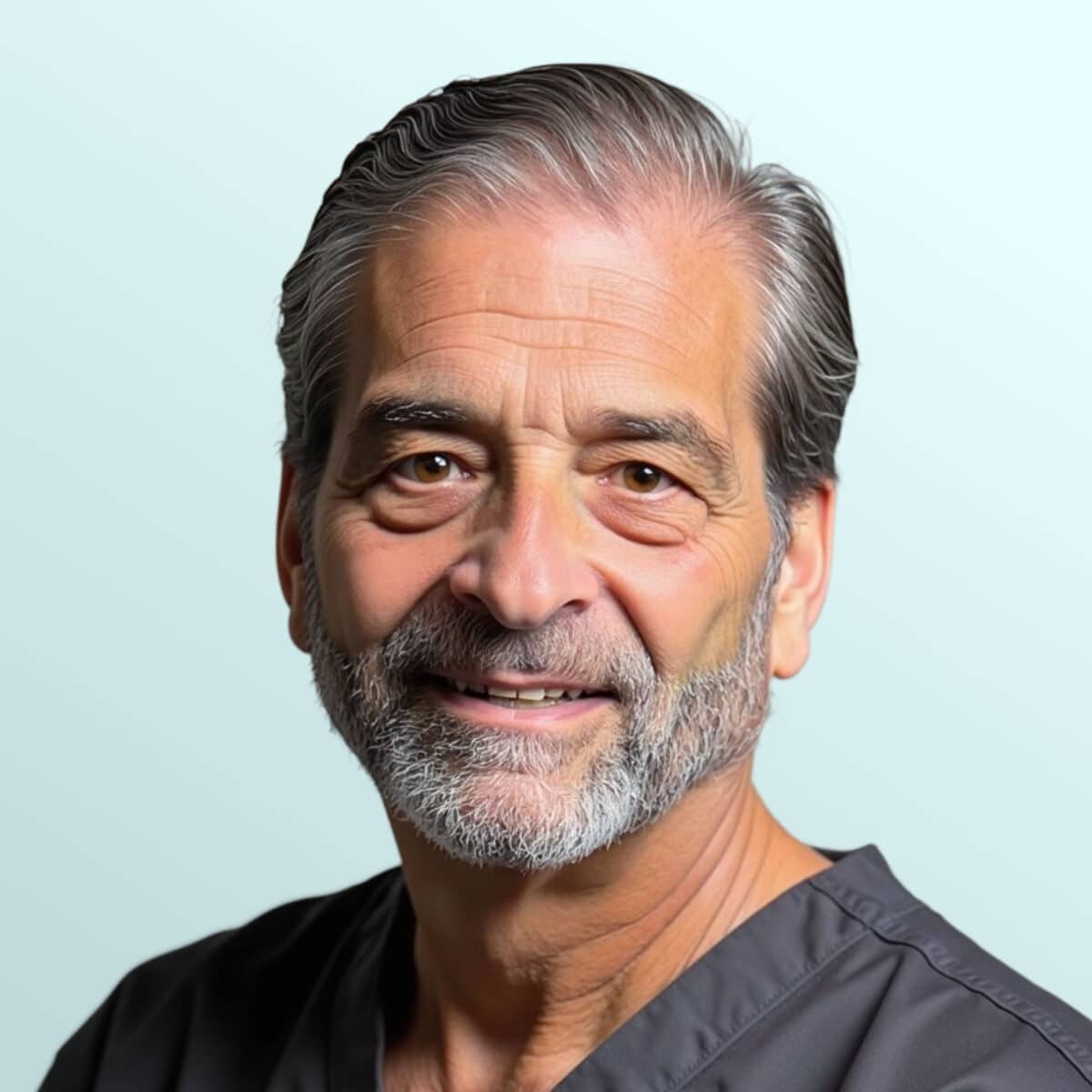 A mature man with gray hair and beard, wearing a navy suit, white shirt, and patterned tie, poses against a light blue background.