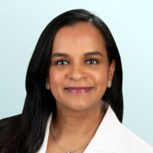 A woman with long dark hair, wearing a white coat, earrings, and a necklace, smiles at the camera against a light blue background.