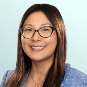 A woman with long brown hair and glasses, wearing a blue blazer and gold necklace, smiles at the camera against a light blue background.