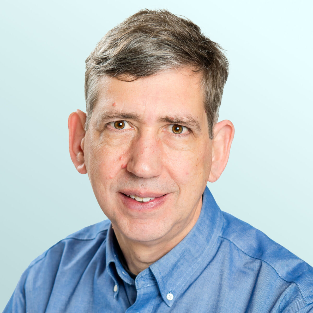 A middle-aged man with short brown hair and fair skin, wearing a blue button-up shirt, smiles gently at the camera against a light blue background.