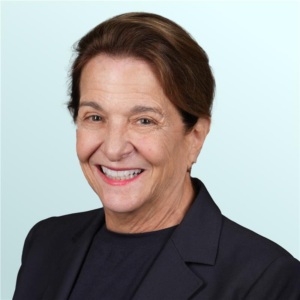 A middle-aged woman with long, light brown hair smiles warmly at the camera. She is wearing a sleeveless black top with beige trim, set against a light blue background.