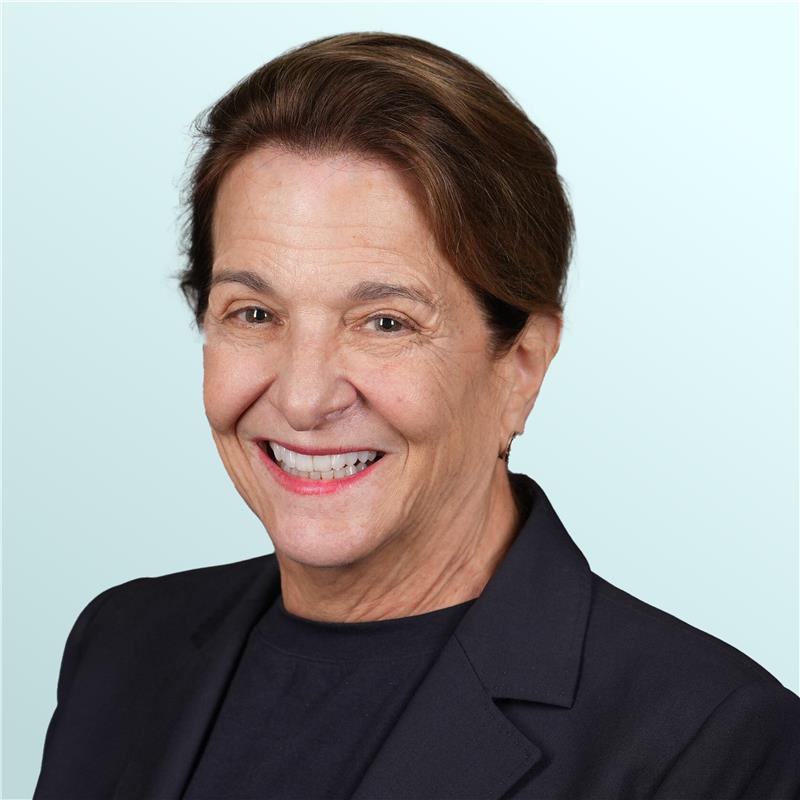 A middle-aged woman with long, light brown hair smiles warmly at the camera. She is wearing a sleeveless black top with beige trim, set against a light blue background.