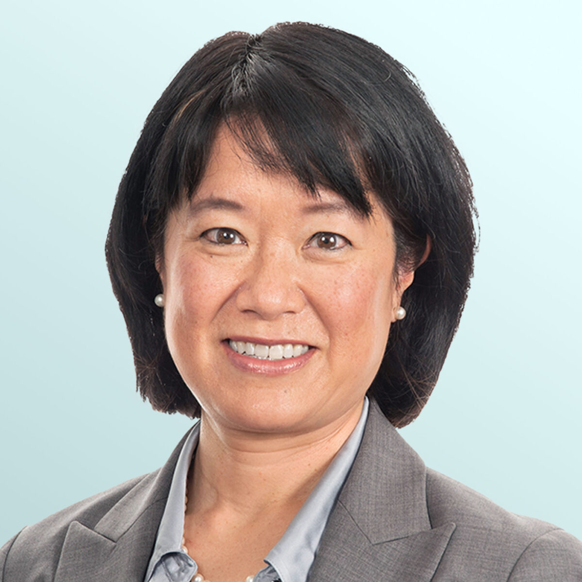 Portrait of an Asian woman with short black hair, wearing a gray blazer, light blue shirt, pearl necklace, and small pearl earrings, smiling in front of a light blue background.