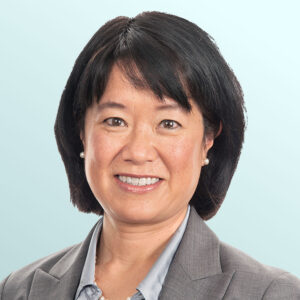 Portrait of an Asian woman with short black hair, wearing a gray blazer, light blue shirt, pearl necklace, and small pearl earrings, smiling in front of a light blue background.