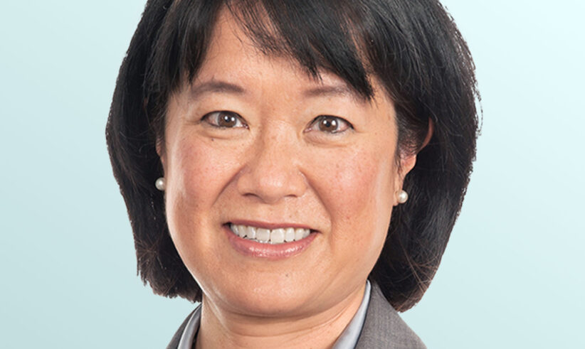 Portrait of an Asian woman with short black hair, wearing a gray blazer, light blue shirt, pearl necklace, and small pearl earrings, smiling in front of a light blue background.