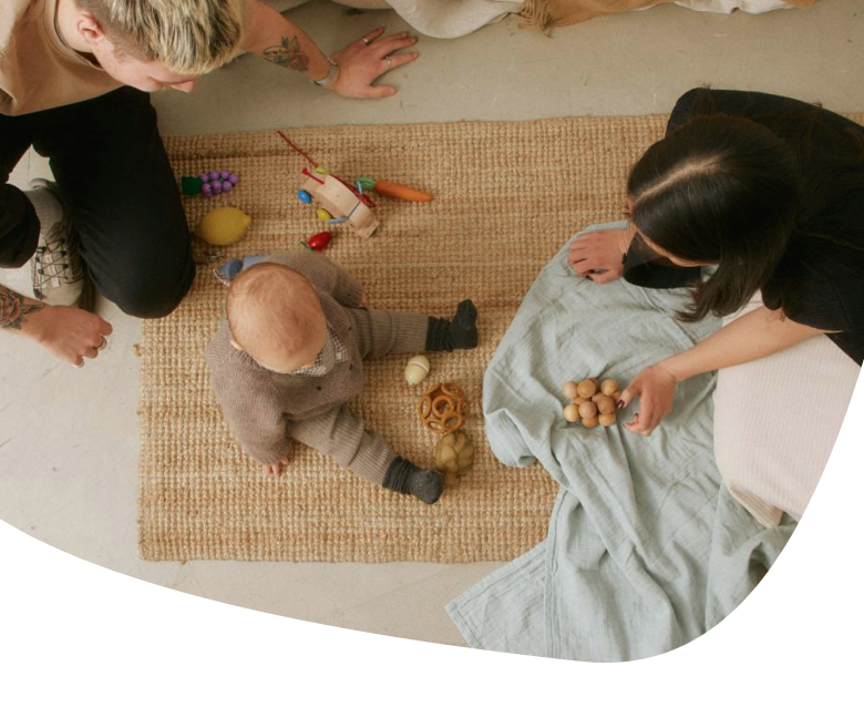 Two adults sit on the floor with a baby on a woven rug. The baby is surrounded by wooden toys and looks up, while one adult holds small objects and a light-colored blanket is spread nearby.