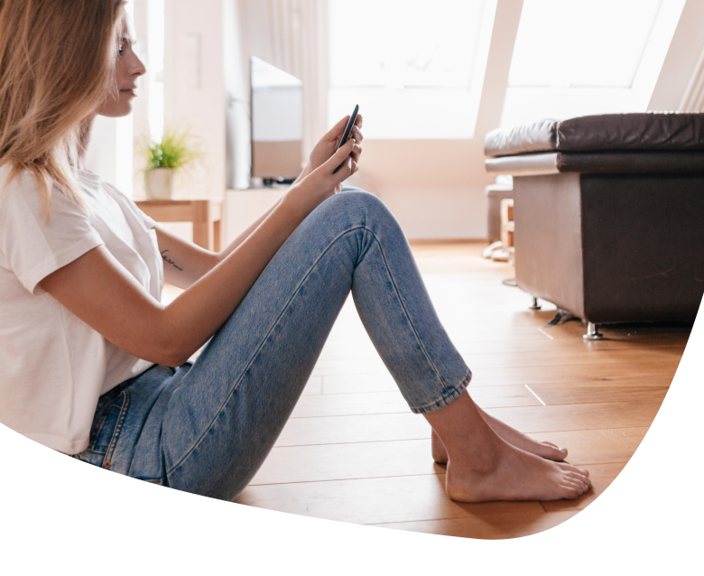 A woman sits barefoot on a wooden floor, leaning against a sofa and looking at her phone. She is wearing a white t-shirt and blue jeans in a bright, modern living room.
