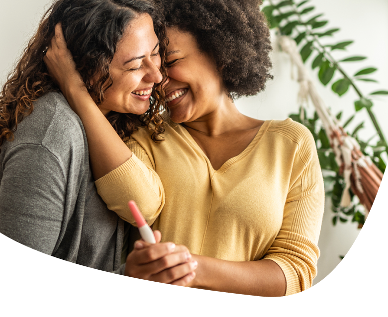 Two women smile and embrace joyfully while holding a positive pregnancy test. Indoor setting with green plants in the background.