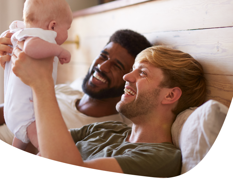 Two men lie on a bed, smiling and looking at a baby one of them is holding up. The scene reflects warmth and happiness in a cozy, home setting.