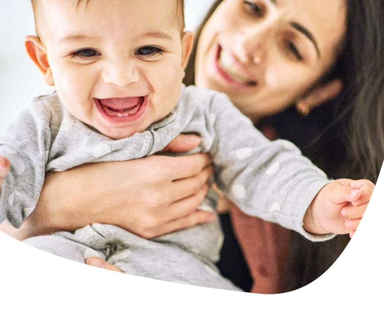 A smiling baby wearing a grey outfit is being held by a woman who is also smiling in the background.