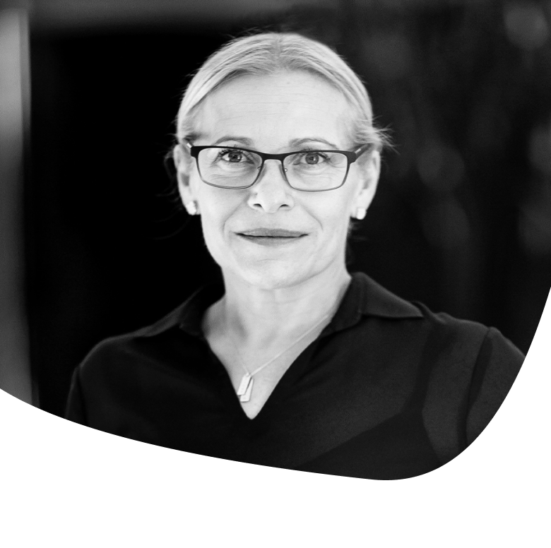 A woman with light hair pulled back, wearing glasses, a dark collared shirt, and a necklace, looks at the camera with a slight smile against a dark, blurred background. The image is in black and white.